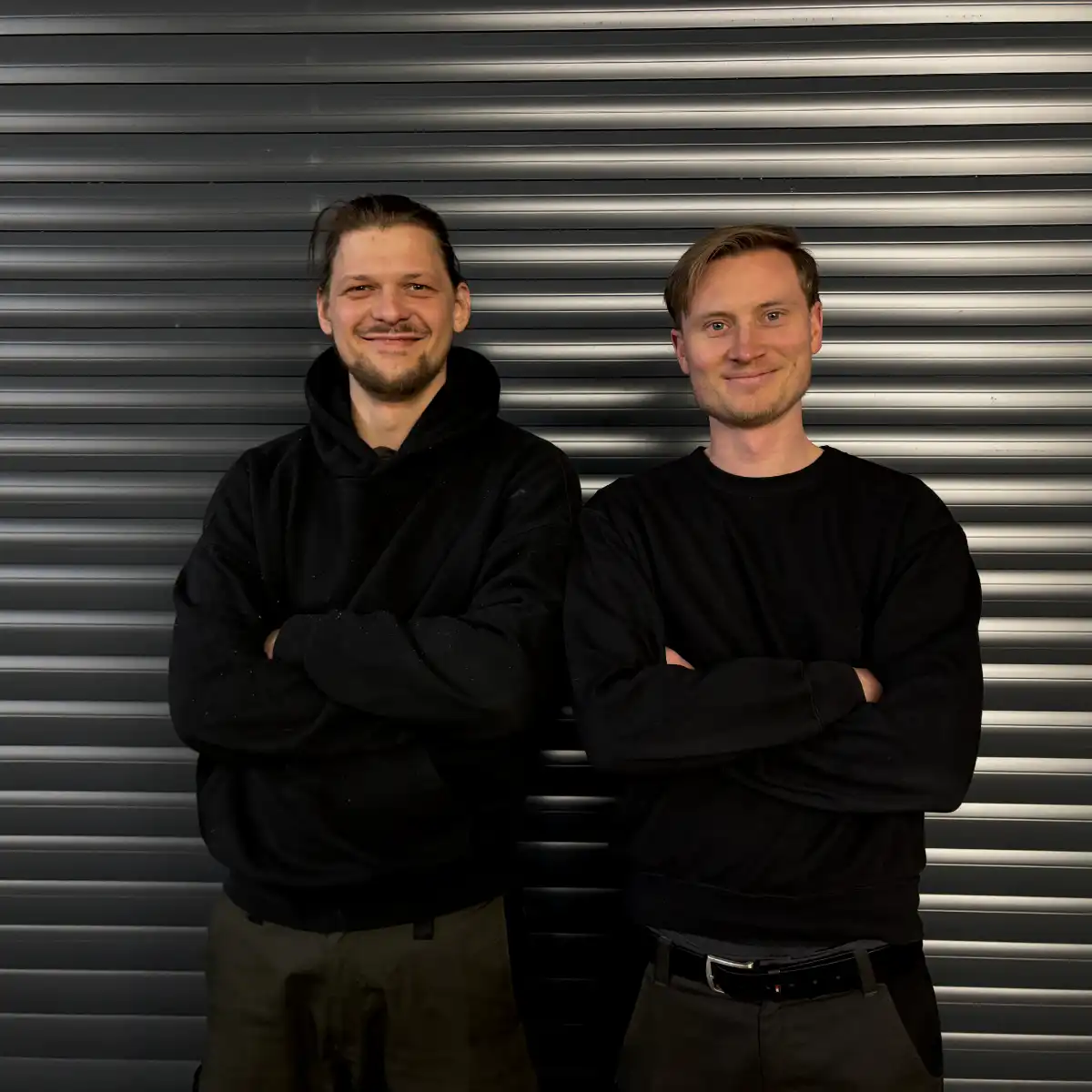 Zwei Männer in schwarzer Kleidung vor Metallwand.
